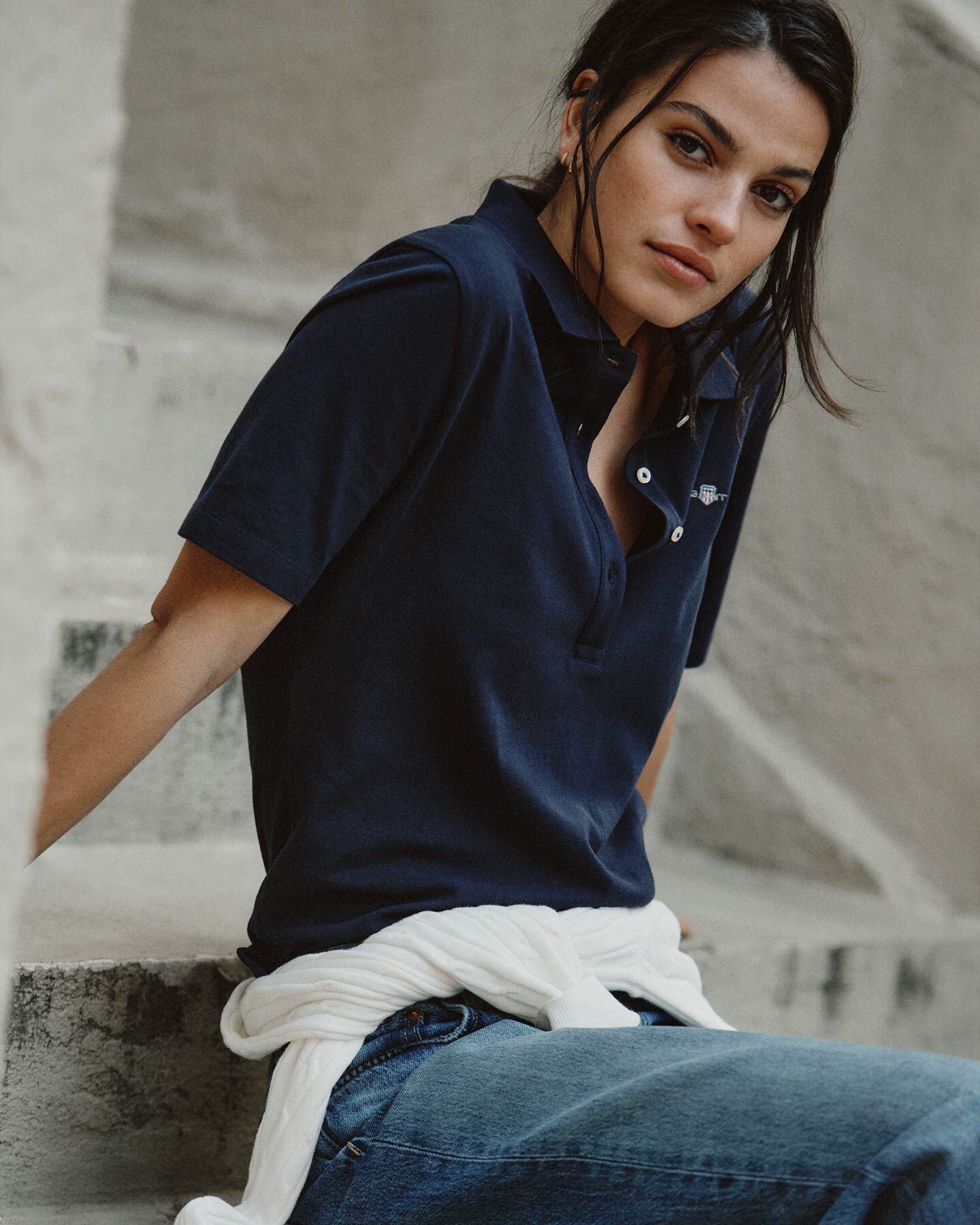 Woman in a navy Gant polo shirt and jeans sits on stone steps.